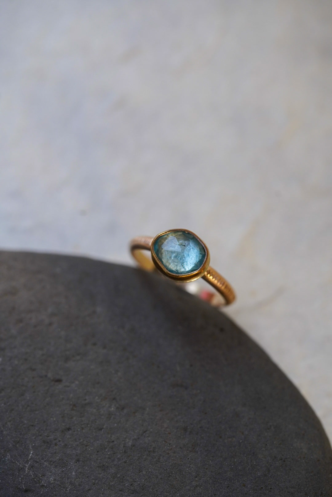 A delicate gold ring with a round light blue gemstone, resting on a smooth dark gray stone against a light beige textured background.