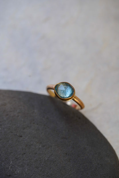 A delicate gold ring with a round light blue gemstone, resting on a smooth dark gray stone against a light beige textured background.