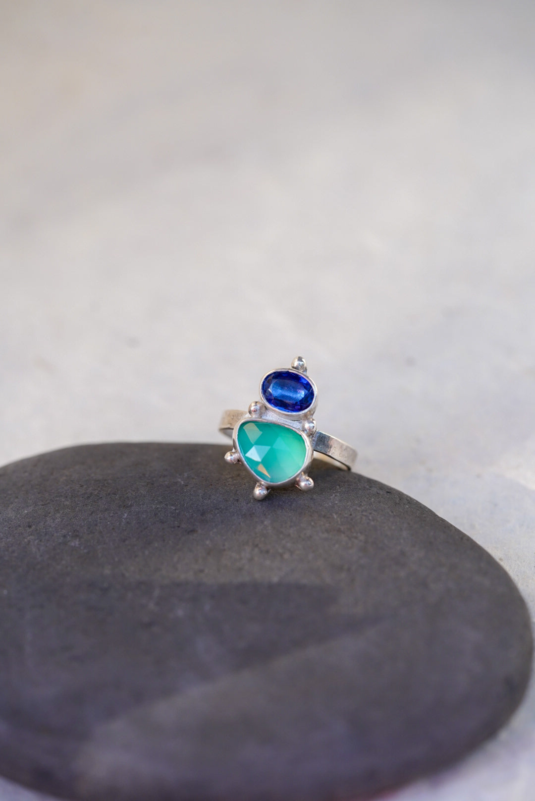 A silver ring featuring a green chalcedony gemstone and a smaller oval dark blue kyanite, displayed on a dark gray stone against a light gray background.