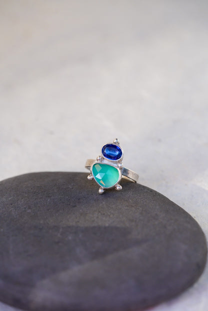 A silver ring featuring a green chalcedony gemstone and a smaller oval dark blue kyanite, displayed on a dark gray stone against a light gray background.