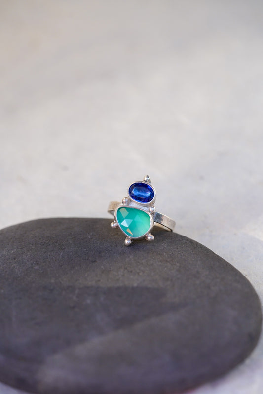 A silver ring featuring a green chalcedony gemstone and a smaller oval dark blue kyanite, displayed on a dark gray stone against a light gray background.