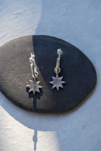 A pair of silver star hoop earrings resting on a dark stone against a light textured background.