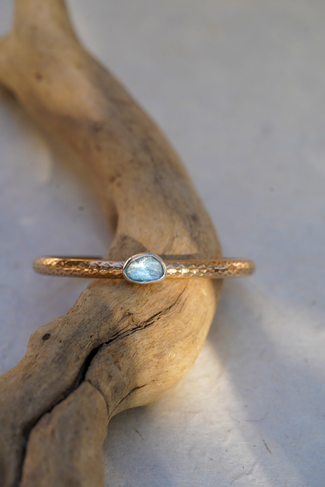 A delicate stamped gold cuff with a small oval light blue aquamarine, resting on a piece of natural driftwood against a light gray background.