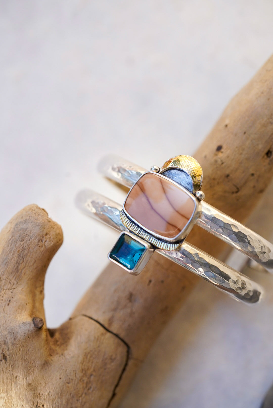 A double-band silver cuff with a central rectangular peach stone, a small blue square gemstone below it, and a textured hammered finish, displayed on driftwood.