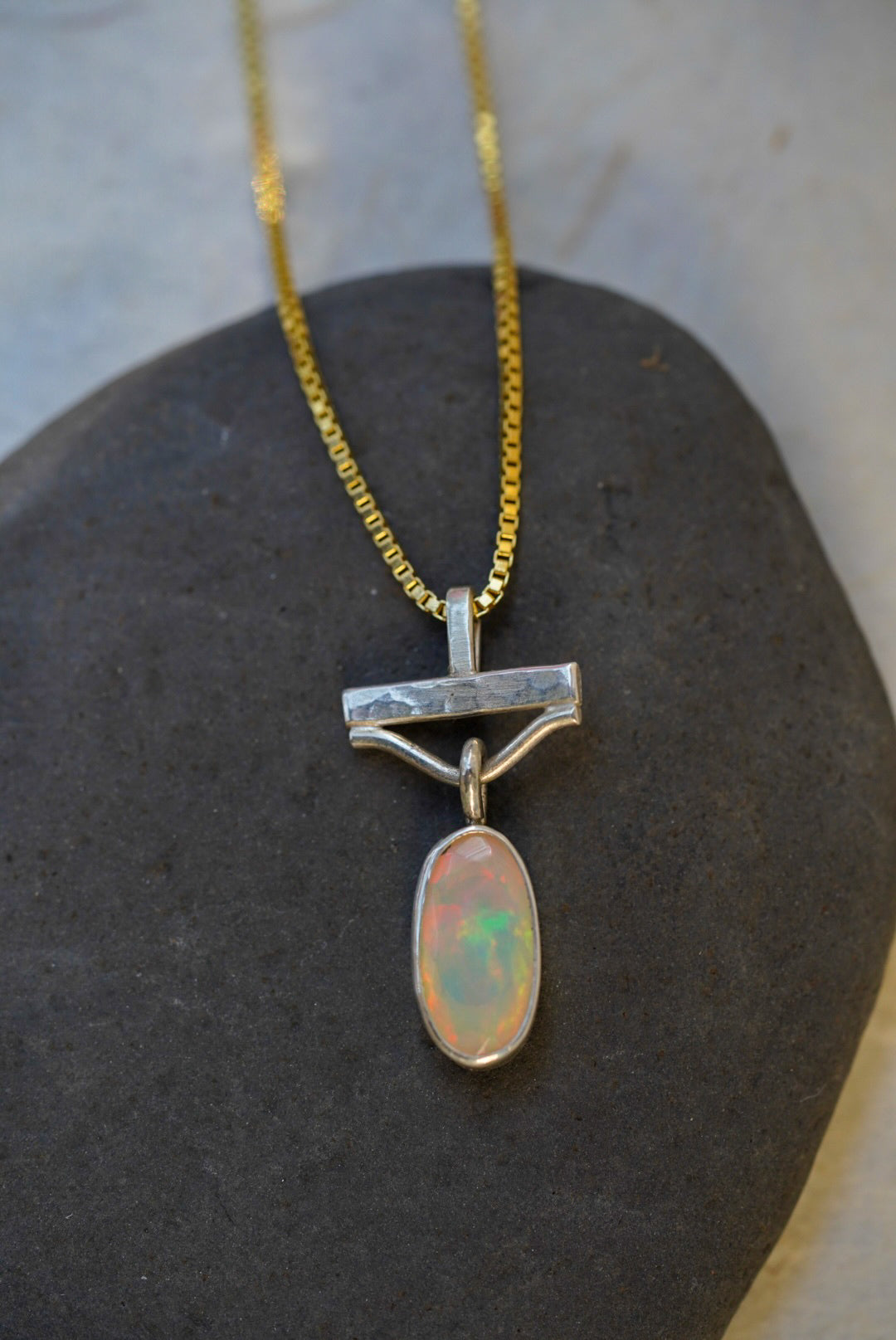 A silver pendant necklace with a hammered bar design and an oval opal stone, displayed on a dark stone surface.
