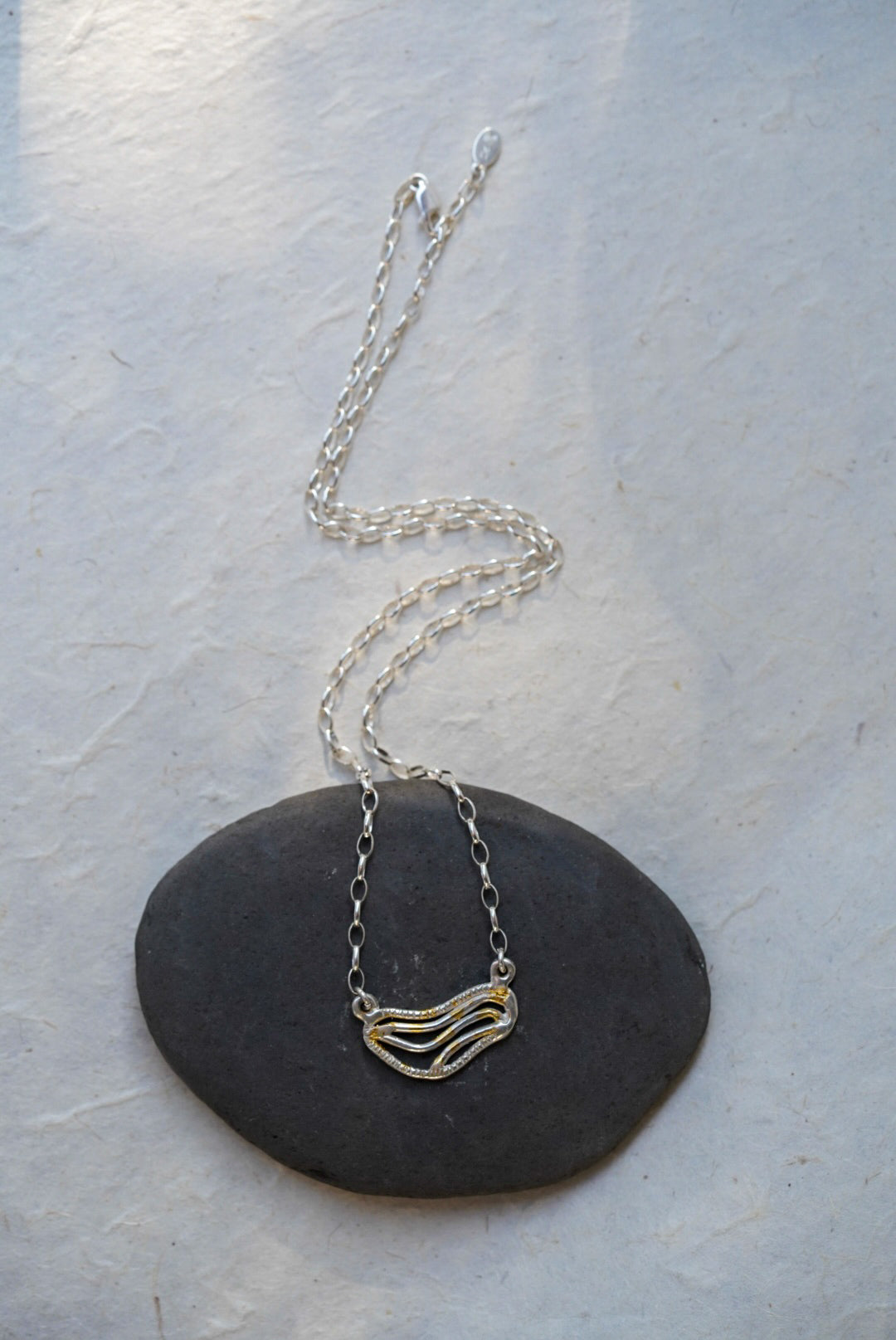 A silver chain necklace with organic shaped pendant featuring textured gold dusting, displayed on a smooth dark gray stone against a light gray background.