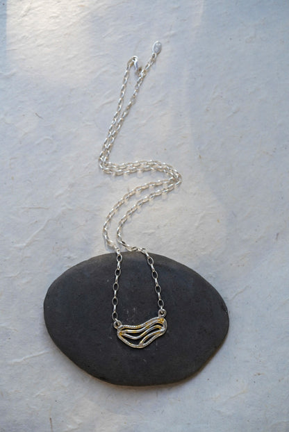 A silver chain necklace with organic shaped pendant featuring textured gold dusting, displayed on a smooth dark gray stone against a light gray background.