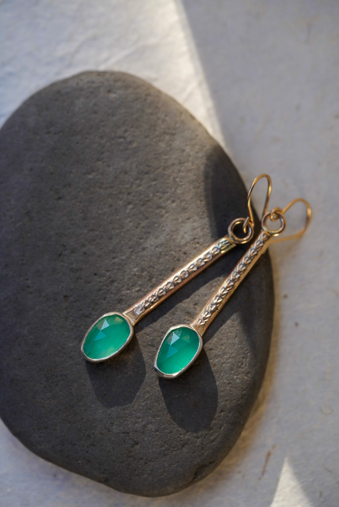 A pair of gold drop earrings with marquise-cut emerald green chalcedony, displayed on a dark gray stone against a light background.