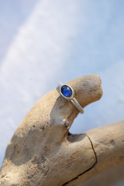 A silver ring with a round blue labradorite gemstone, displayed on a piece of driftwood against a soft, out-of-focus light background.