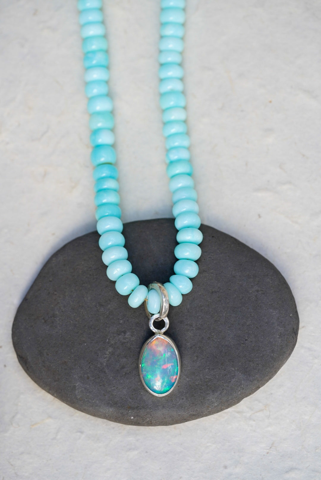 A baby blue beaded necklace with removable charm in rainbow opal, set upon a black rock on a light background.