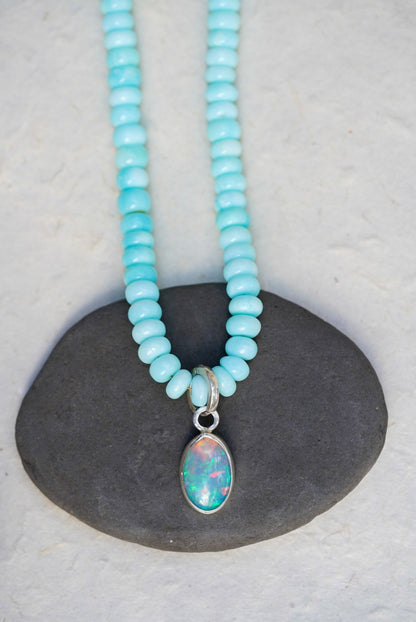 A baby blue beaded necklace with removable charm in rainbow opal, set upon a black rock on a light background.
