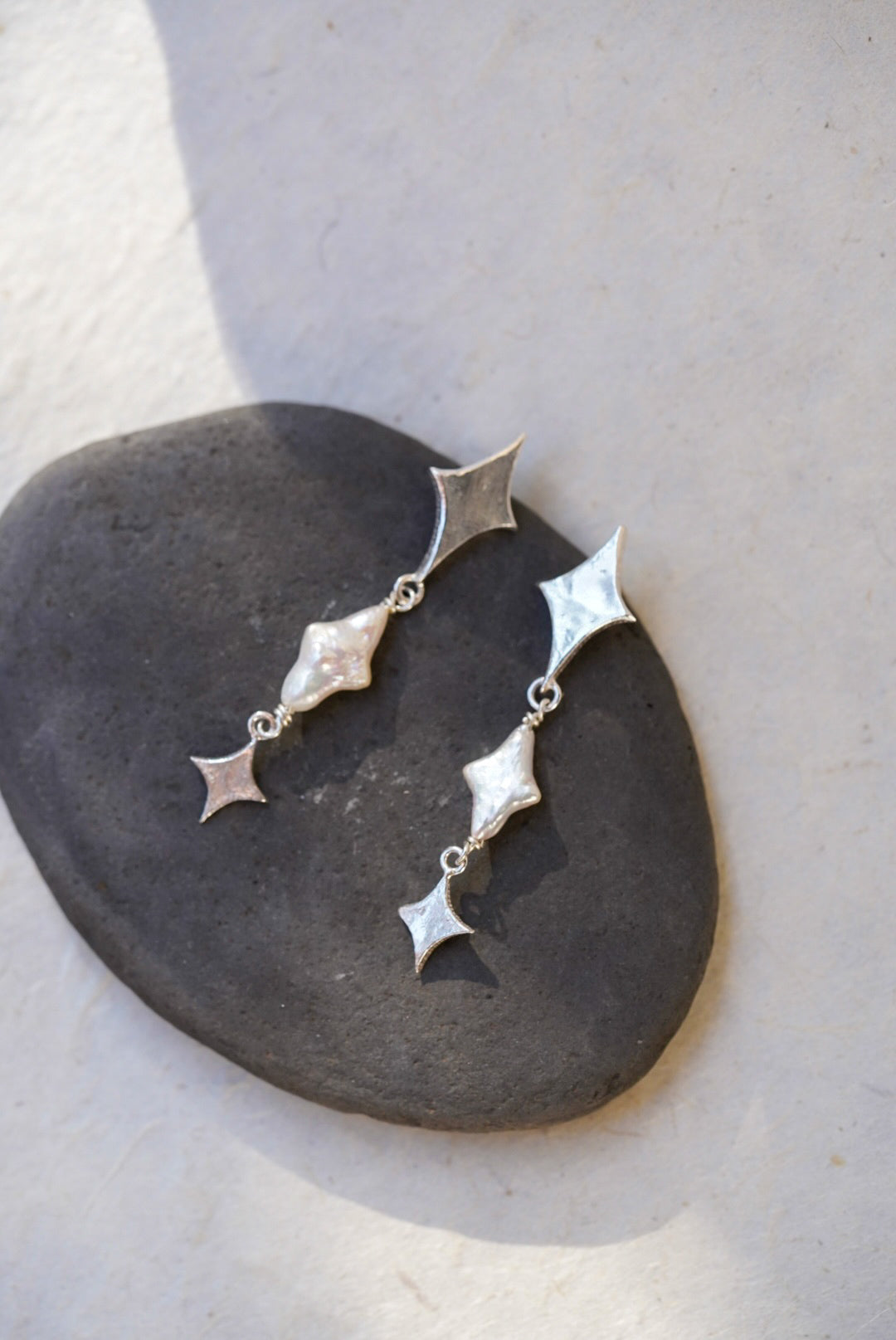 A pair of silver dangle earrings with star-shaped and star pearls, displayed on a dark stone against a light beige background.