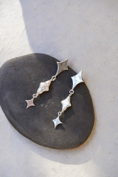 A pair of silver dangle earrings with star-shaped and star pearls, displayed on a dark stone against a light beige background.