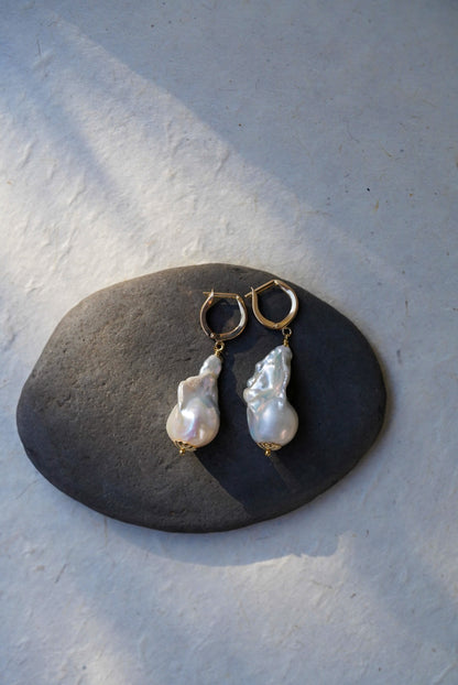 A pair of gold hoop earrings with irregular white baroque pearl drops, displayed on a dark gray stone against a textured light gray background.