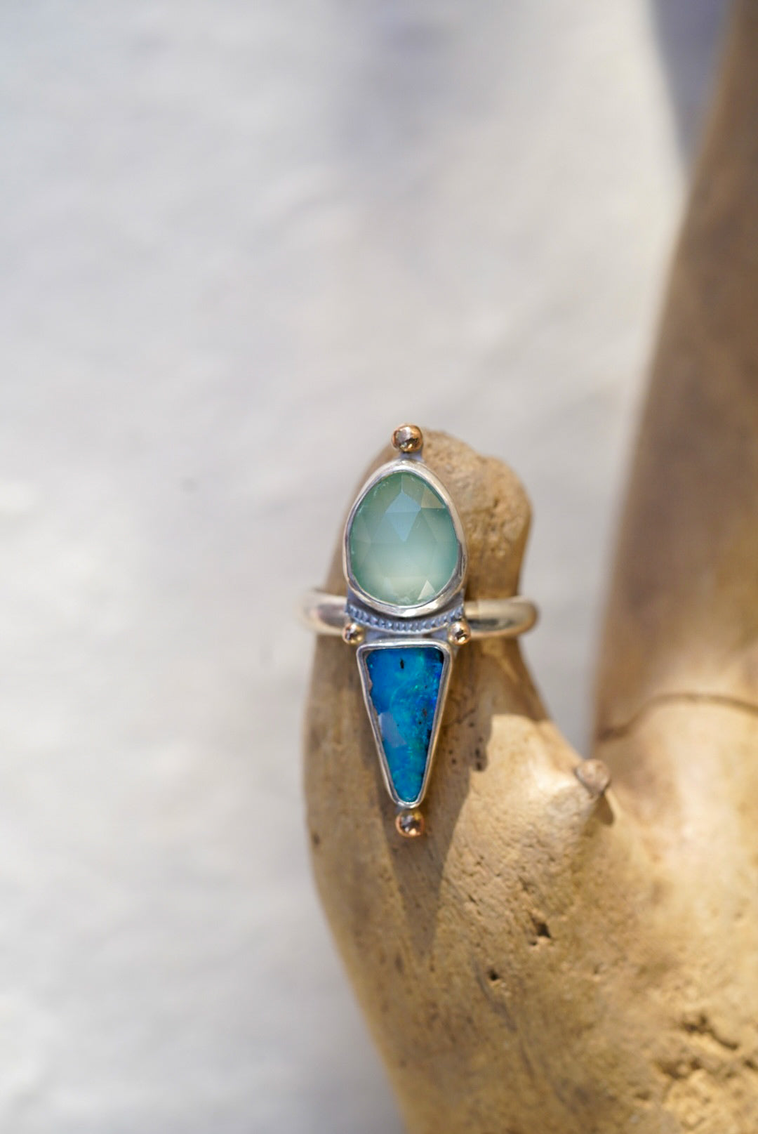 A silver ring with a pale blue stone and a triangular bright blue opal stone, displayed on a textured tan stone stand.