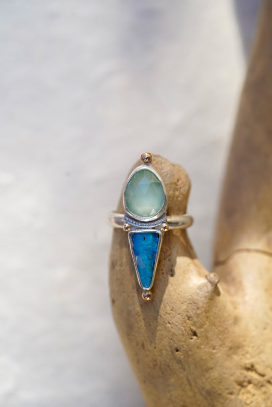 A silver ring with a pale blue stone and a triangular bright blue opal stone, displayed on a textured tan stone stand.