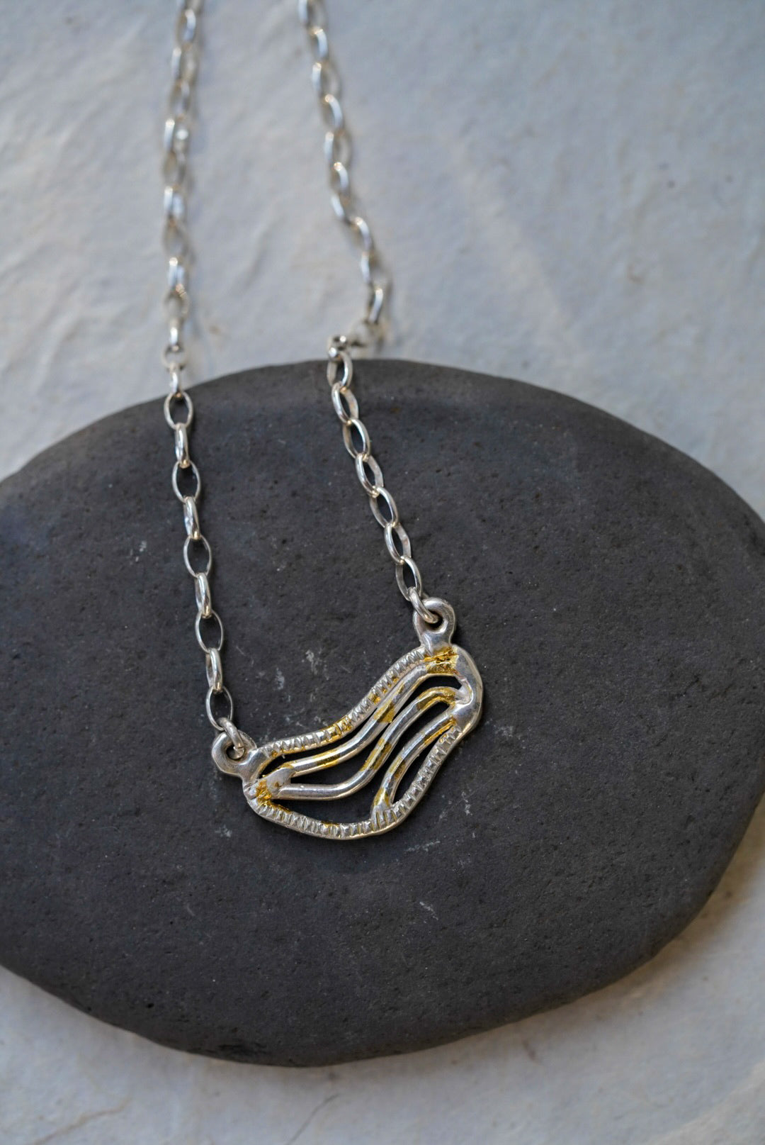 A silver chain necklace with organic shaped pendant featuring textured gold dusting, displayed on a smooth dark gray stone against a light gray background.