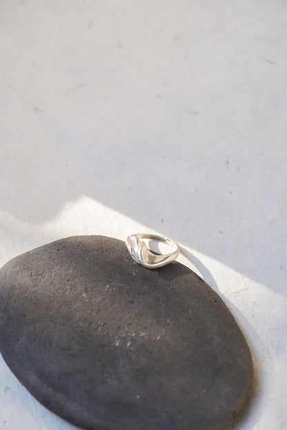 A polished silver ring with a curved, organic shape resting on a smooth dark gray stone against a light gray background.