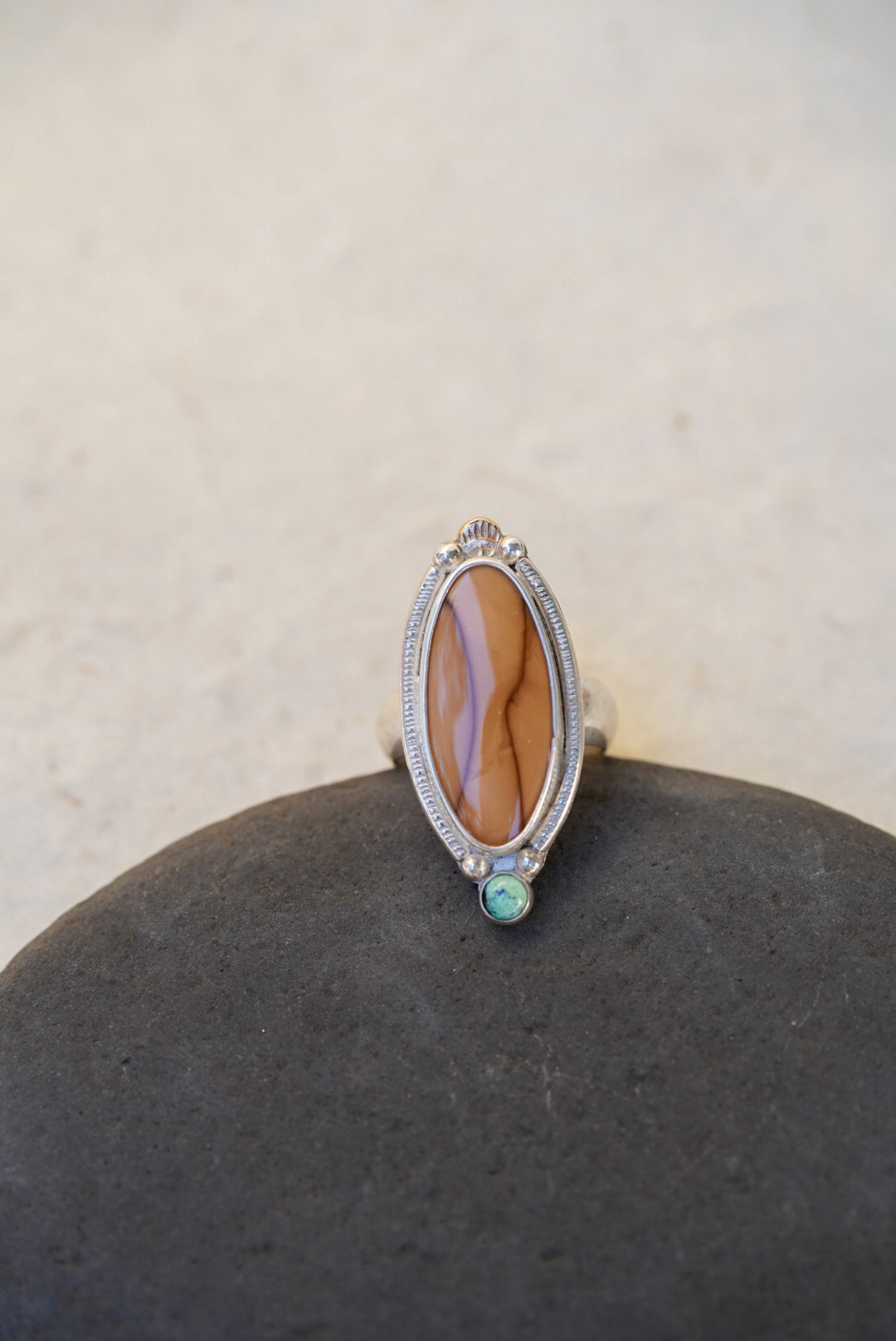 A silver ring with a large brown stone and a small green accent stone, displayed on a dark stone surface against a light beige background.