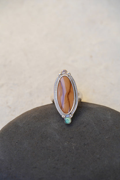 A silver ring with a large brown stone and a small green accent stone, displayed on a dark stone surface against a light beige background.