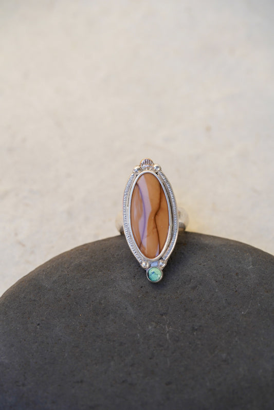 A silver ring with a large brown stone and a small green accent stone, displayed on a dark stone surface against a light beige background.