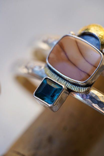 A close-up of a double-band silver cuff with a central rectangular peach stone, a small blue square gemstone below it, and a textured hammered finish, displayed on driftwood.