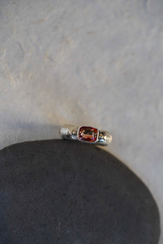 A silver ring with a rectangular red-pink gemstone, displayed on a dark gray stone against a light gray textured background.