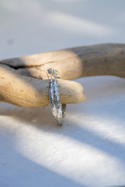 A pair of silver hoop earrings with intricate detailing, resting on a piece of driftwood over a white surface with soft shadows.