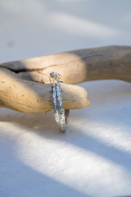 A pair of silver hoop earrings with intricate detailing, resting on a piece of driftwood over a white surface with soft shadows.