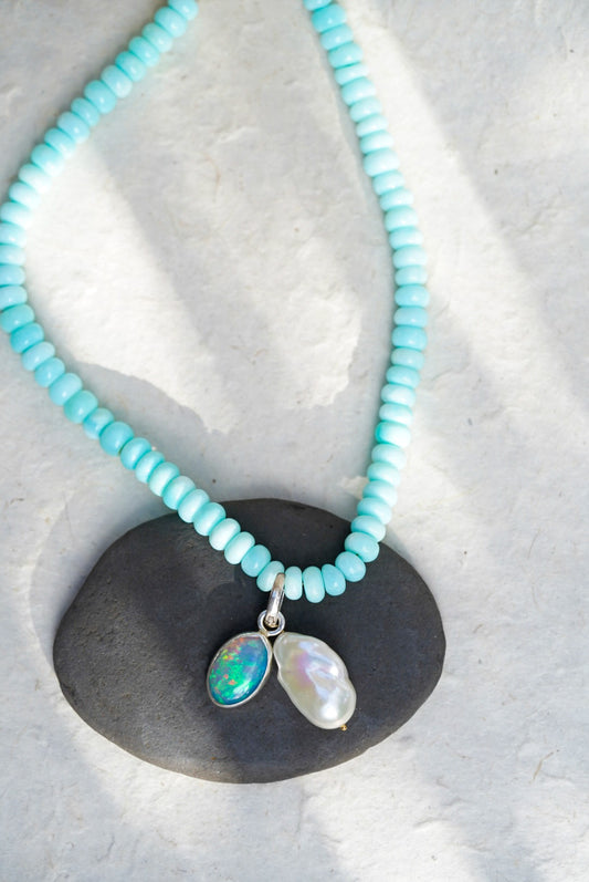 A baby blue beaded necklace with removable charms in rainbow opal and large baroque pearl, set upon a black rock on a light background.