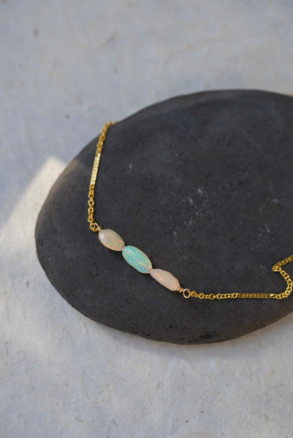A delicate gold chain necklace with three opal  oval beads, including two light pink and one pale blue-green bead, displayed on a dark stone against a light textured background.