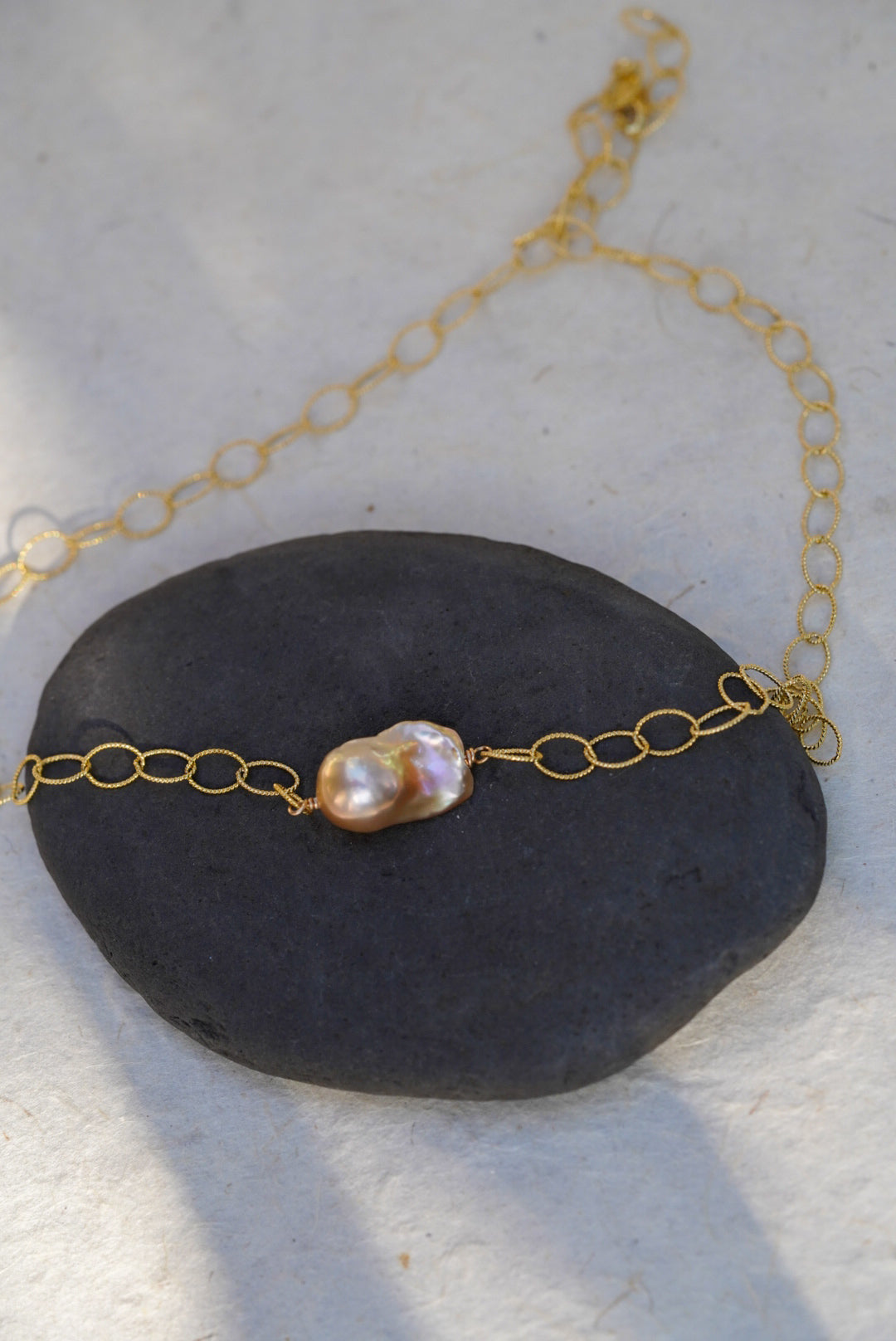 A delicate gold chain necklace featuring a single baroque pearl pendant, displayed on a dark gray stone against a light textured background.