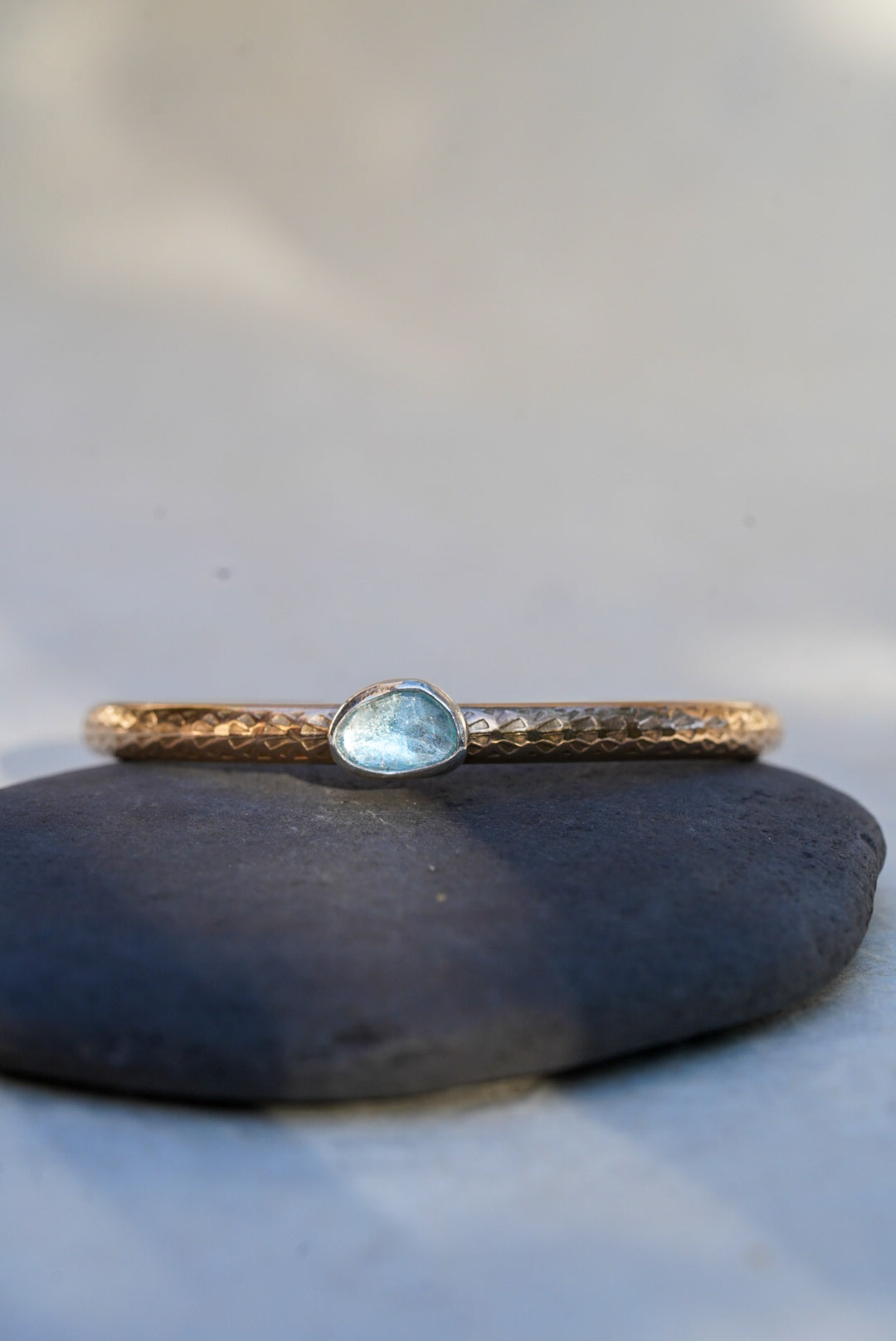 A delicate hammered gold bangle bracelet with a small, irregular-shaped light blue gemstone, resting on a smooth dark gray stone against a soft, light gray background.