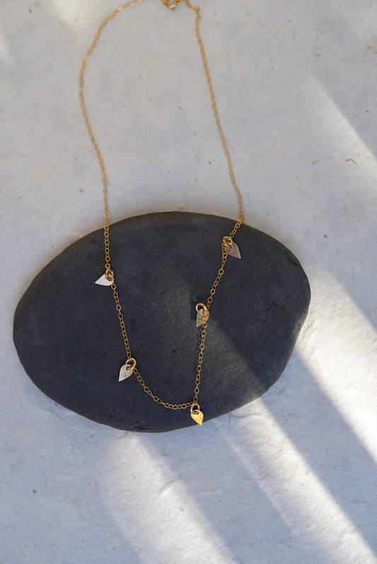 A delicate gold necklace with small charms, displayed on a dark stone over a white textured surface with soft shadows.