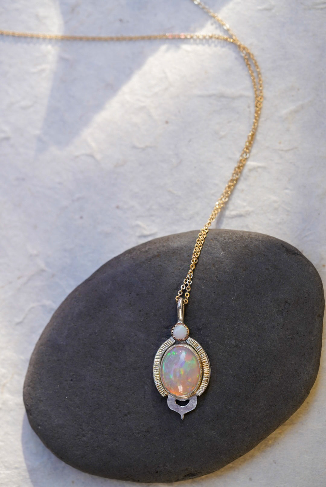 A gold chain necklace with an oval iridescent opal pendant and a small white opal, displayed on a dark stone surface.