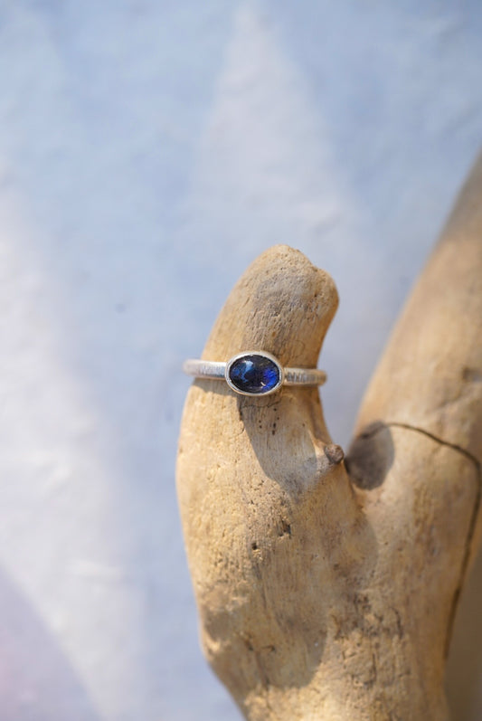 A silver ring with a round blue labradorite gemstone, displayed on a piece of driftwood against a soft, out-of-focus light background.