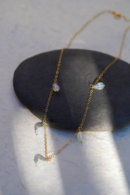 A delicate gold chain necklace with five small, teardrop-shaped iridescent opal beads, draped over a smooth dark gray stone on a textured white surface.