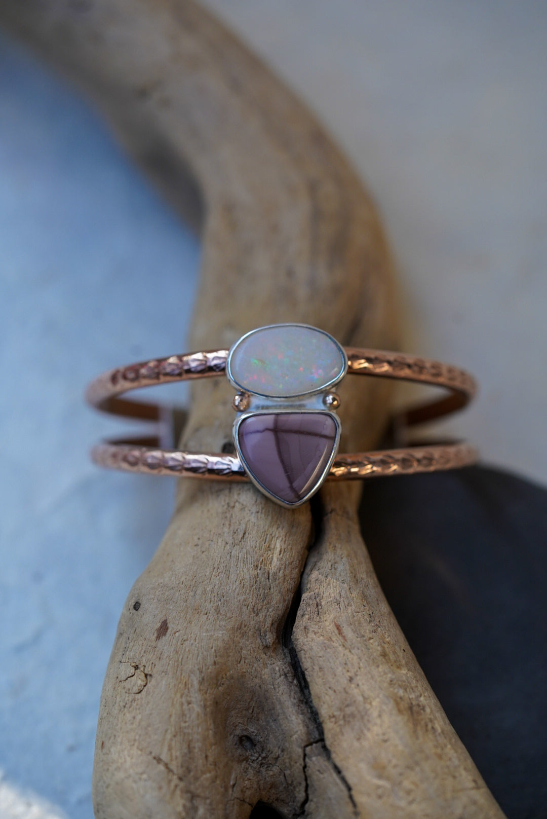 A textured rose gold double band cuff with an opal and jasper, displayed on a piece of driftwood.