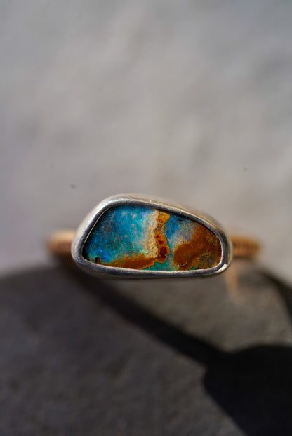 A close-up shot of a handmade ring featuring a irregularly shaped turquoise and rust-colored opal set in a silver bezel, resting on a dark gray stone with a soft-focus light gray background.