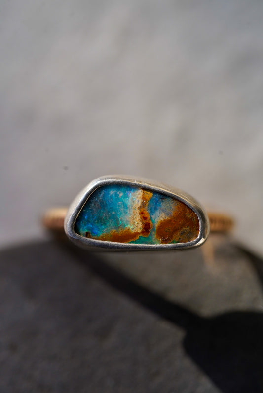 A close-up shot of a handmade ring featuring a irregularly shaped turquoise and rust-colored opal set in a silver bezel, resting on a dark gray stone with a soft-focus light gray background.