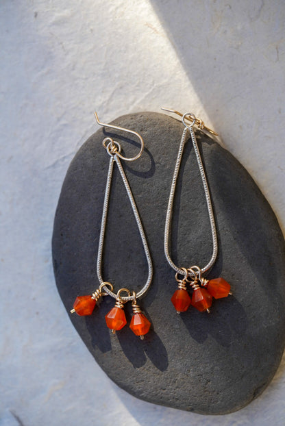 A pair of long dangle earrings with silver teardrop shape  and small orange faceted carnelian beads, displayed on a dark stone against a light textured background.
