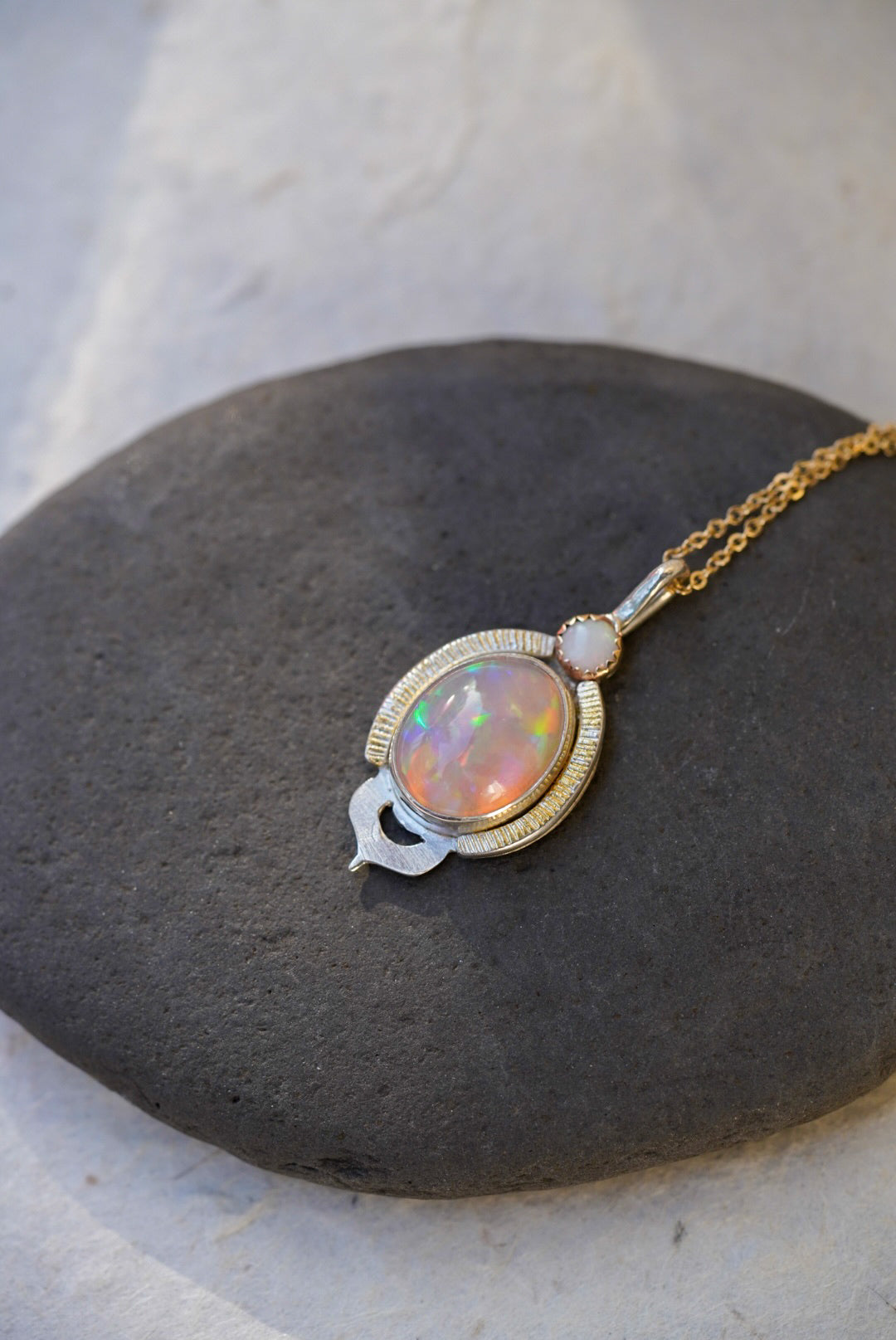 A gold and silver pendant necklace featuring a colorful opal stone, resting on a dark stone slab against a light textured background.