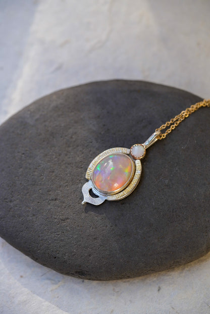 A gold and silver pendant necklace featuring a colorful opal stone, resting on a dark stone slab against a light textured background.