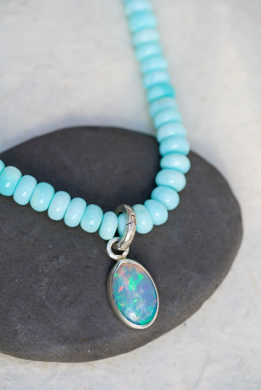 A baby blue beaded necklace with removable charm in rainbow opal, set upon a black rock on a light background.