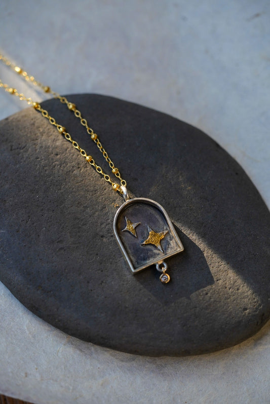 A gold chain necklace with a arched pendant containing two silver stars with a dusting of gold, resting on a dark stone against a textured light background.