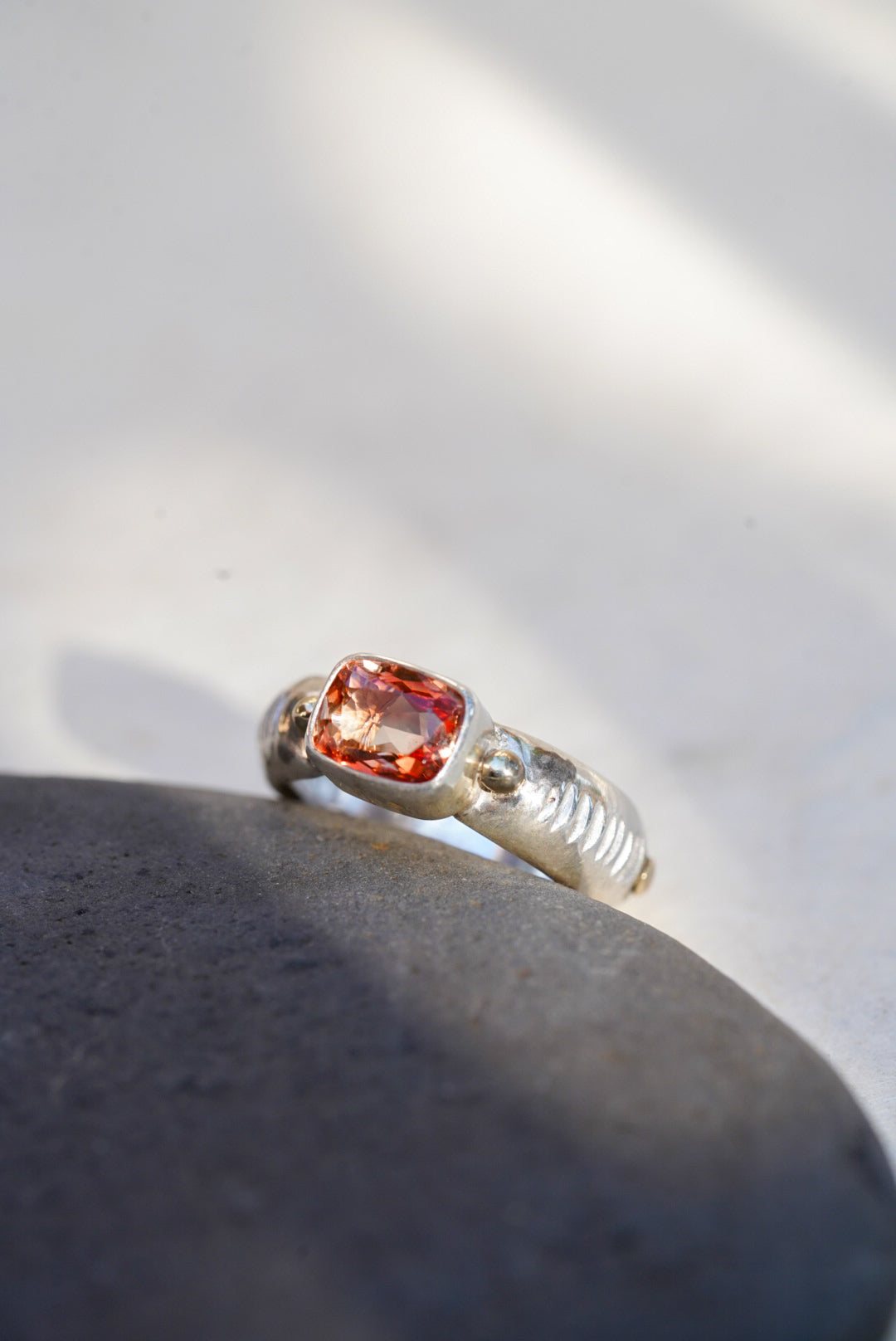 A silver ring with a cushion-cut orange gemstone, resting on a dark gray stone against a bright white background with soft shadows.