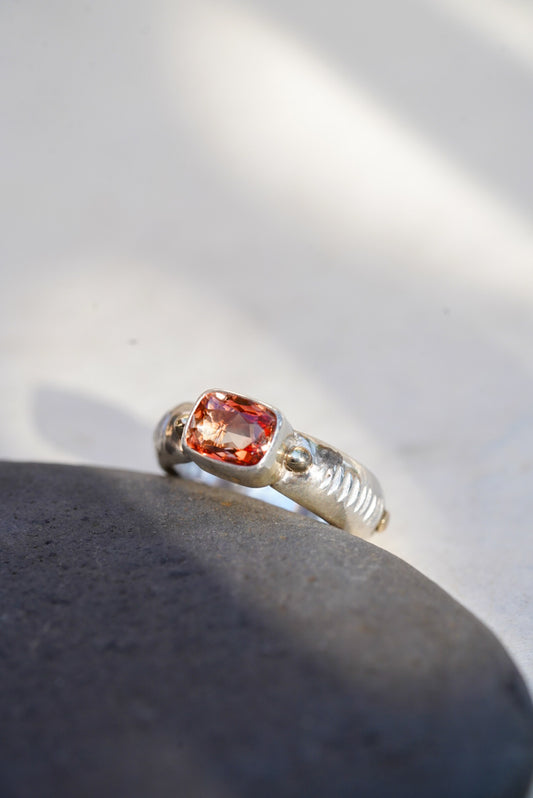 A silver ring with a cushion-cut orange gemstone, resting on a dark gray stone against a bright white background with soft shadows.