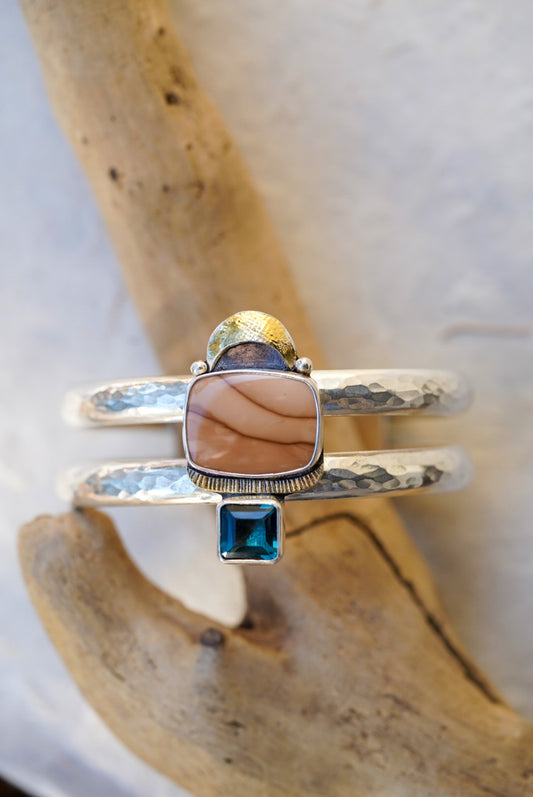 A double-band silver cuff with a central rectangular peach stone, a small blue square gemstone below it, and a textured hammered finish, displayed on driftwood.