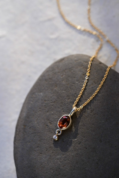A delicate gold chain necklace with an oval amber-brown gemstone pendant and a small diamond accent stone, displayed on a dark gray stone slab against a light beige background.