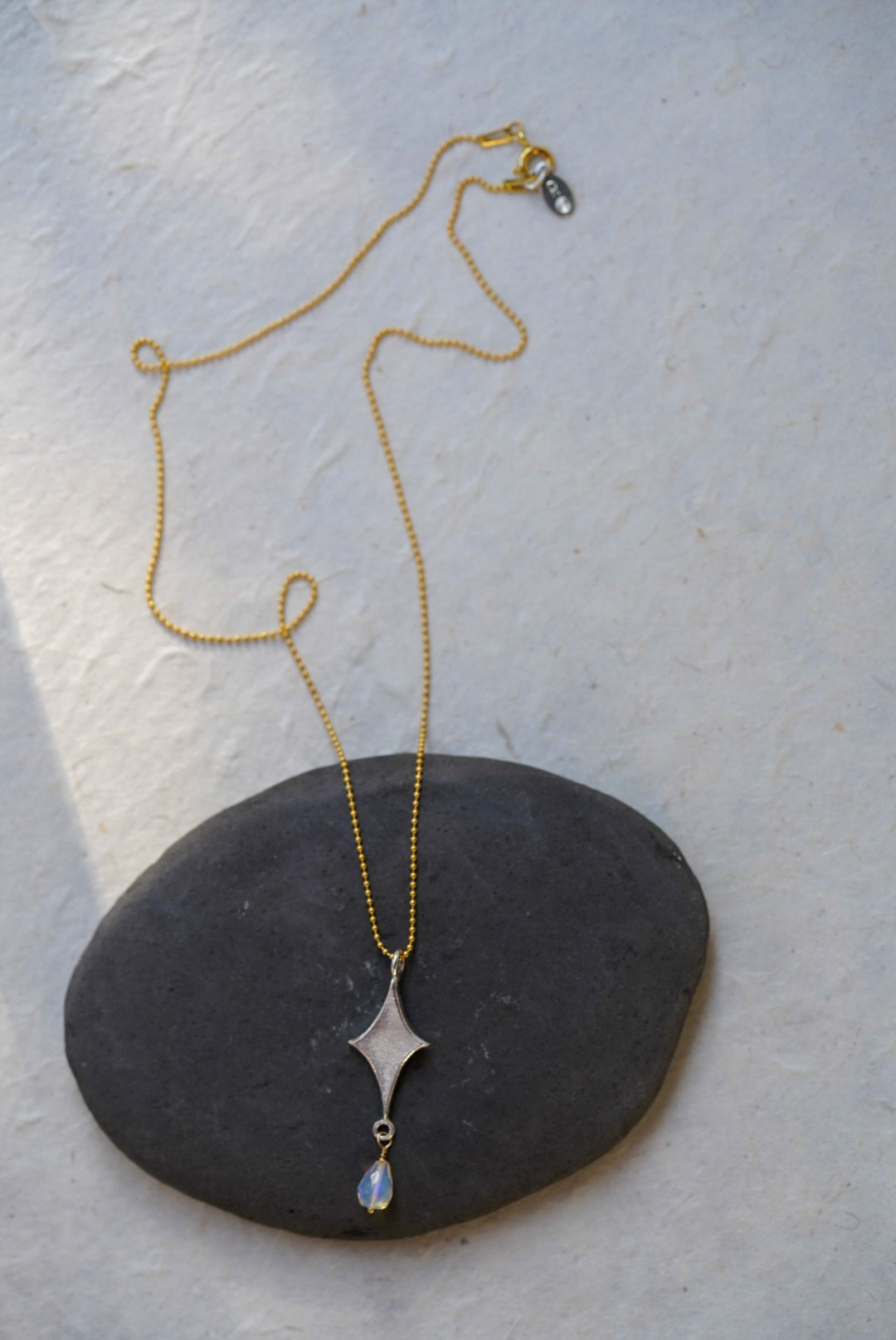 A delicate gold beaded chain necklace with a silver starburst pendant and a small teardrop opal, displayed on a dark stone over a textured off-white background.